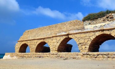 Lionsgate Travel introduces the ancient Roman aqueduct in Caesarea Maritima, featuring long arches along a sandy beach under a blue sky.